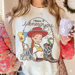 May include: A white t-shirt featuring a graphic of a cowgirl with red hair, a red hat, and a yellow vest. The design includes a blue balloon, a horseshoe, blocks labeled 13, T, and S, a cowboy boot with a green snake, and the text I knew it.