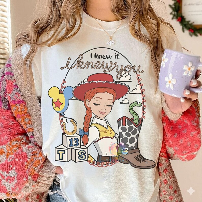 May include: A white t-shirt featuring a graphic of a cowgirl with red hair, a red hat, and a yellow vest. The design includes a blue balloon, a horseshoe, blocks labeled 13, T, and S, a cowboy boot with a green snake, and the text I knew it.