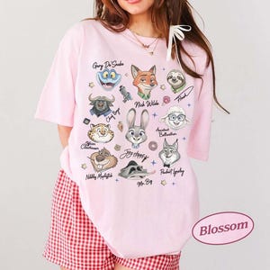 K&ouml;nnte beinhalten: Hellrosa T-Shirt mit Zeichentrickfiguren aus Zootopia, darunter Judy Hopps und Nick Wilde. Das Shirt hat das Wort "Blossom" in einem rosa Oval unten rechts.