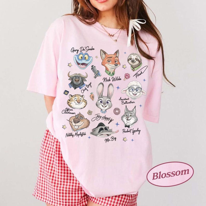 K&ouml;nnte beinhalten: Hellrosa T-Shirt mit Zeichentrickfiguren aus Zootopia, darunter Judy Hopps und Nick Wilde. Das Shirt hat das Wort "Blossom" in einem rosa Oval unten rechts.