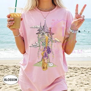 May include: Pink t-shirt featuring a cartoon illustration of a tower with a long-haired character hanging upside down. The design includes the text "OH HI!" in a speech bubble. The word "BLOSSOM" is printed on the bottom left.