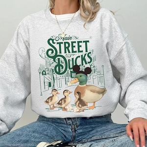 Op de afbeelding: Een lichtgrijze sweatshirt met een afbeelding van een eend met Mickey Mouse-oren en drie eendenkuikens. De tekst "Main Street Ducks" is in het groen gedrukt, met een grillig ontwerp. De sweatshirt is een casual stijl.