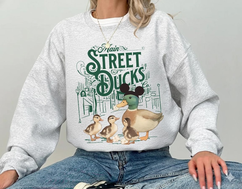 Op de afbeelding: Een lichtgrijze sweatshirt met een afbeelding van een eend met Mickey Mouse-oren en drie eendenkuikens. De tekst "Main Street Ducks" is in het groen gedrukt, met een grillig ontwerp. De sweatshirt is een casual stijl.