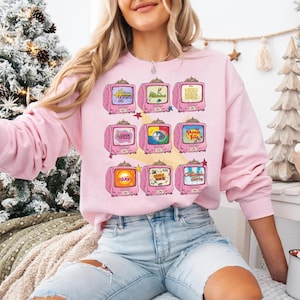 May include: A light pink sweatshirt featuring a repeating pattern of pink retro televisions. Each TV screen displays a different cartoon image. The sweatshirt has long sleeves and a crew neck. The model is wearing light blue jeans.