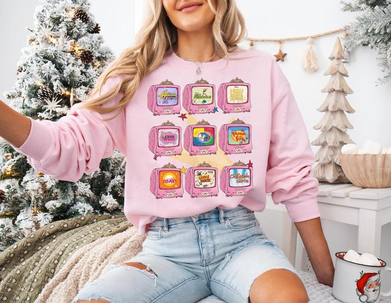 May include: A light pink sweatshirt featuring a repeating pattern of pink retro televisions. Each TV screen displays a different cartoon image. The sweatshirt has long sleeves and a crew neck. The model is wearing light blue jeans.