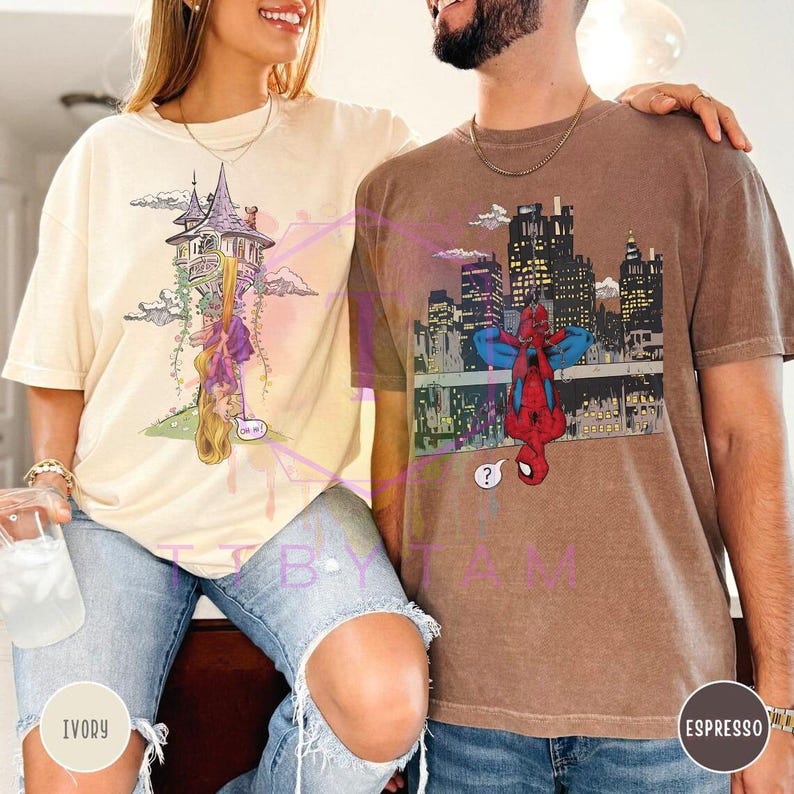 May include: Ivory and espresso-colored t-shirts. The ivory shirt features a cartoon of a tower with a princess hanging from her hair. The espresso shirt has a Spider-Man graphic with a city skyline background.