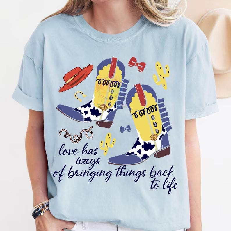 Pu&ograve; includere: Una t-shirt azzurra con una stampa grafica di due stivali da cowboy gialli e blu con dettagli maculati, un cappello rosso, due cactus gialli, una corda e piccoli fiocchi rossi e blu. Il testo recita, Love has ways of bringing things back to life.