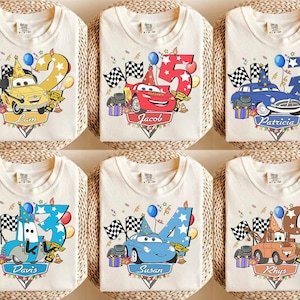 May include: Cream-colored t-shirts featuring colorful cartoon car designs with numbers and names. Each shirt has a different car character, number, and name, with racing flags and party decorations. Perfect for a birthday celebration.
