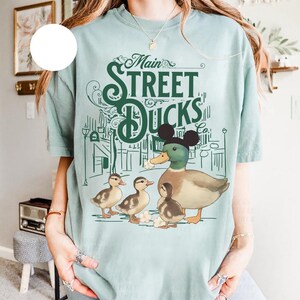 Op de afbeelding: Lichtblauw T-shirt met een afbeelding van een eend met Mickey Mouse oren en drie eendenkuikens. De tekst "Main Street Ducks Co." staat boven de eendenfamilie gedrukt, met een stadsgezichtontwerp. Het shirt is gemaakt van een zacht, comfortabel materiaal.