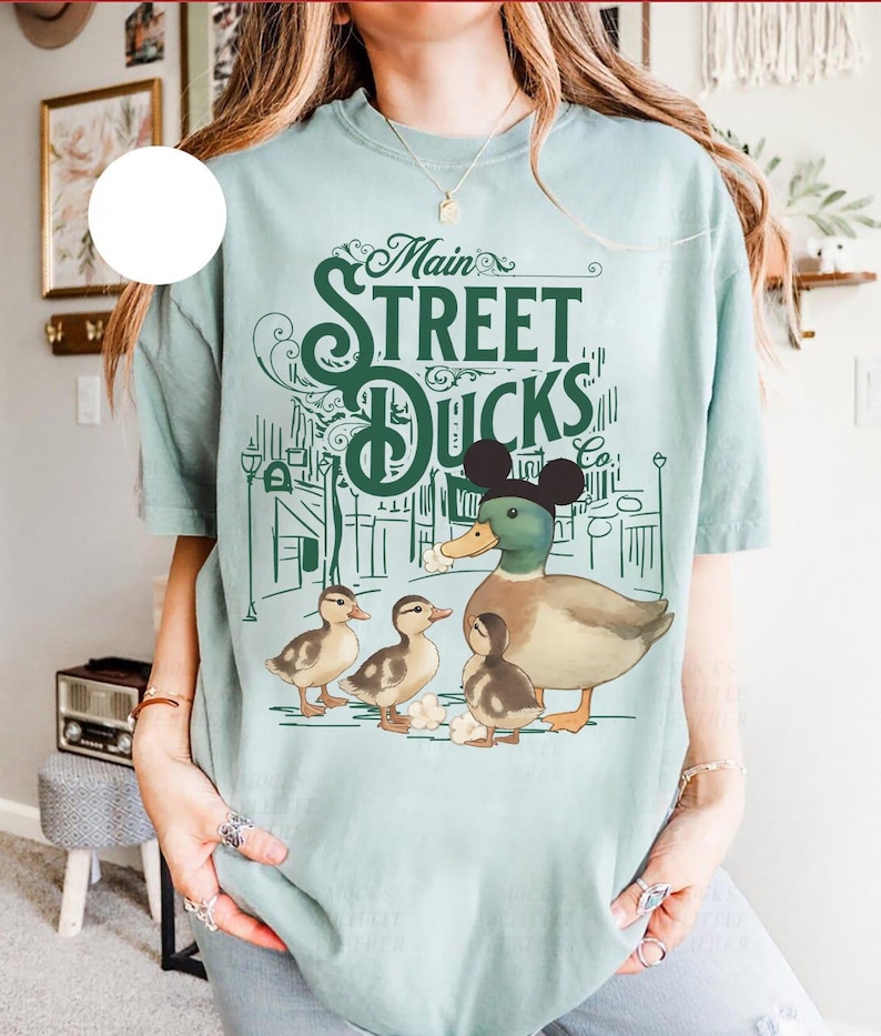 Op de afbeelding: Lichtblauw T-shirt met een afbeelding van een eend met Mickey Mouse oren en drie eendenkuikens. De tekst "Main Street Ducks Co." staat boven de eendenfamilie gedrukt, met een stadsgezichtontwerp. Het shirt is gemaakt van een zacht, comfortabel materiaal.