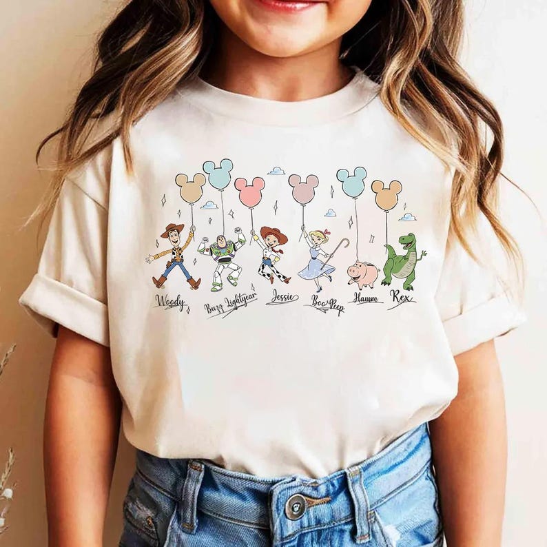 May include: A cream-colored t-shirt featuring cartoon characters from the Toy Story movie. The characters are holding Mickey Mouse-shaped balloons. The names of the characters are written below them.