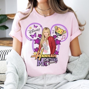 May include: A light pink t-shirt featuring a graphic of Hannah Montana. The design includes the Disney logo, stars, butterflies, musical notes, and the text "20 Years". The graphic also features a picture of Hannah Montana with a red jacket and a guitar.
