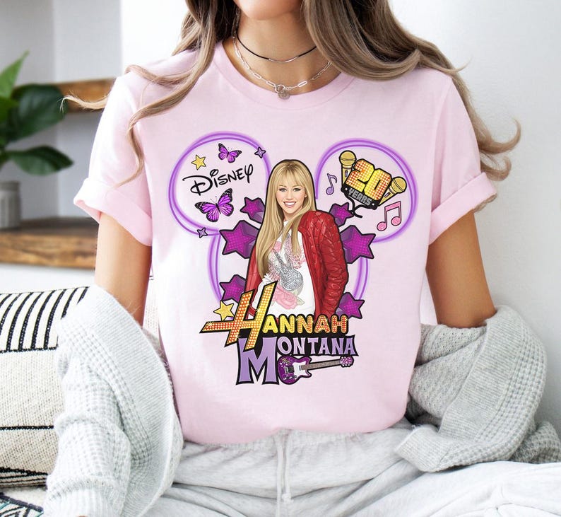 May include: A light pink t-shirt featuring a graphic of Hannah Montana. The design includes the Disney logo, stars, butterflies, musical notes, and the text "20 Years". The graphic also features a picture of Hannah Montana with a red jacket and a guitar.
