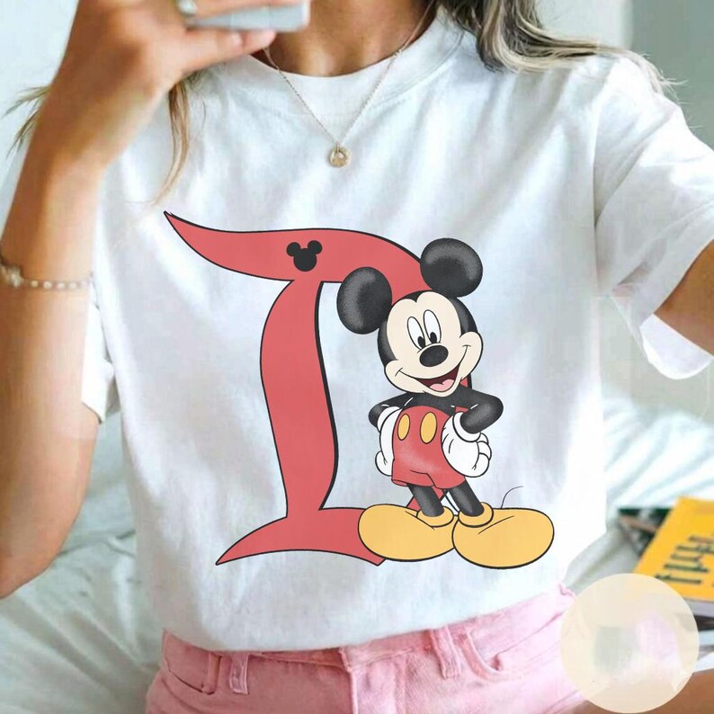 Peut inclure: Un t-shirt blanc &agrave; manches courtes arborant un grand D rouge avec une petite silhouette de t&ecirc;te de Mickey Mouse noire dans la courbe sup&eacute;rieure. &Agrave; c&ocirc;t&eacute; de la lettre se trouve une illustration en couleur de Mickey en short rouge et chaussures jaunes.