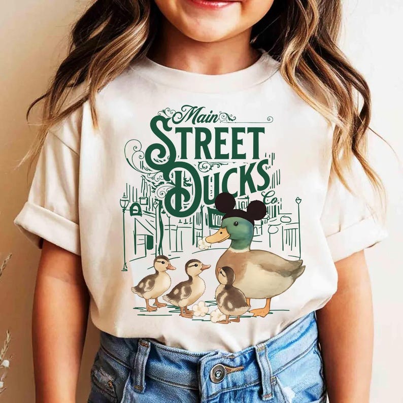Op de afbeelding: Cr&egrave;mekleurig T-shirt met een groene afbeelding van een eendenfamilie en de tekst "Main Street Ducks Co.". De eend in het midden draagt een zwarte Mickey Mouse orenhoed. Het ontwerp bevat een straatbeeld met lantaarnpalen.