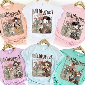 May include: Assorted pastel-colored t-shirts featuring a Wild West design with cartoon characters. Each shirt displays a different character, including Mickey Mouse, Pluto, Donald Duck, and Goofy, in cowboy attire, set against a desert backdrop with cacti.