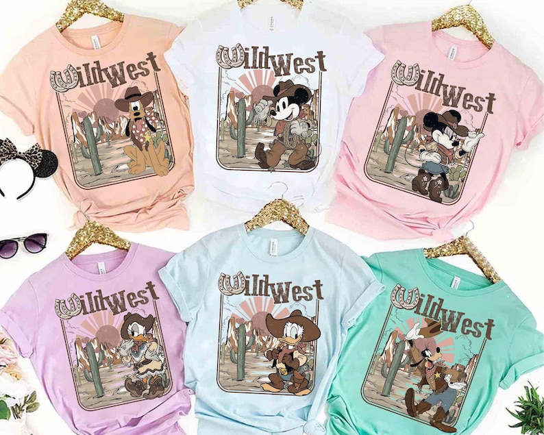 May include: Assorted pastel-colored t-shirts featuring a Wild West design with cartoon characters. Each shirt displays a different character, including Mickey Mouse, Pluto, Donald Duck, and Goofy, in cowboy attire, set against a desert backdrop with cacti.