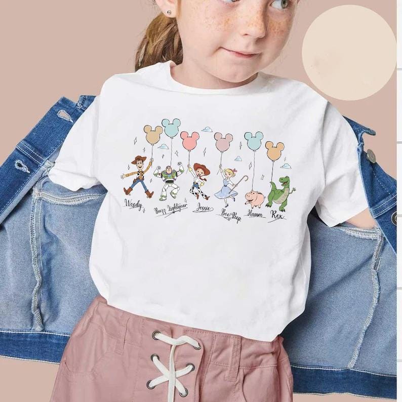May include: White t-shirt featuring a colorful graphic of Toy Story characters holding Mickey Mouse-shaped balloons. The characters include Woody, Buzz Lightyear, Jessie, Bo Peep, Hamm, and Rex. The names of the characters are written below them in a playful font.