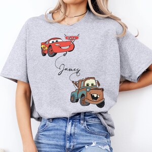 May include: A light gray t-shirt featuring cartoon images of Lightning McQueen and Mater from the movie Cars. The name "James" is written in cursive below Lightning McQueen. The shirt is a short-sleeved crew neck.