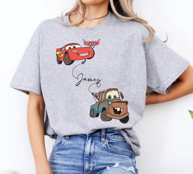 May include: A light gray t-shirt featuring cartoon images of Lightning McQueen and Mater from the movie Cars. The name "James" is written in cursive below Lightning McQueen. The shirt is a short-sleeved crew neck.