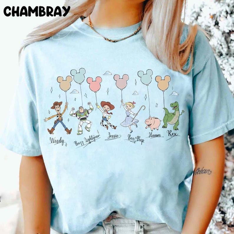May include: Light blue t-shirt featuring a cartoon illustration of characters from a popular animated movie. The characters are holding Mickey Mouse-shaped balloons. The word "CHAMBRAY" is printed at the top.