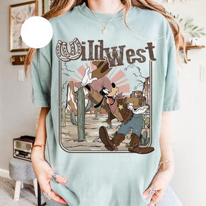 May include: A light blue t-shirt featuring a Wild West design with a cartoon character in cowboy attire. The graphic includes a horseshoe, cactus, and the text "Wild West" in a vintage style. The shirt has short sleeves.
