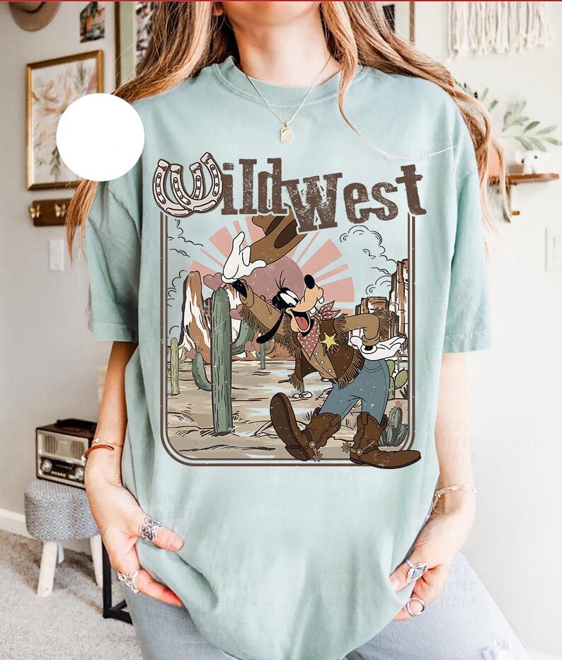 May include: A light blue t-shirt featuring a Wild West design with a cartoon character in cowboy attire. The graphic includes a horseshoe, cactus, and the text "Wild West" in a vintage style. The shirt has short sleeves.