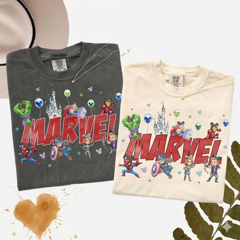 May include: Two t-shirts, one dark gray and one cream, featuring a Marvel design. The design includes Marvel characters, a castle, balloons, and the word "MARVEL" in red. Both shirts have a crew neck.