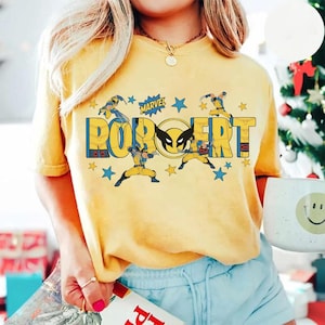 May include: Yellow t-shirt featuring the name "ROBERT" in blue and yellow block letters, with Wolverine graphics and the Marvel logo. The shirt has a crew neck and short sleeves. The design includes stars.