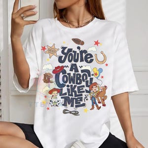 Pu&ograve; includere: Una t-shirt bianca a maniche corte con una stampa grafica recante il testo "You're A Cowboy Like Me". Il design include illustrazioni di una stella da sceriffo, un ferro di cavallo, un lazo, uno stivale e personaggi di Toy Story, tra cui Woody.
