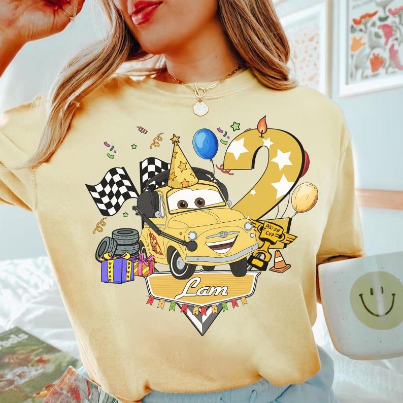 May include: Yellow t-shirt featuring a cartoon car with a party hat, checkered flags, and the number 2. The design includes balloons, confetti, and the name "Lam" on a banner. The shirt is a light yellow color.