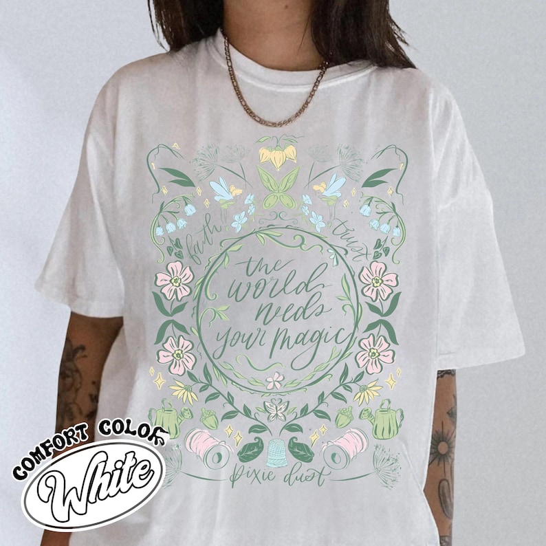 Pu&ograve; includere: T-shirt bianca Comfort Colors con un design floreale e la scritta "the world needs your magic". Il design include fiori rosa e blu, foglie verdi e le parole "faith" e "trust".