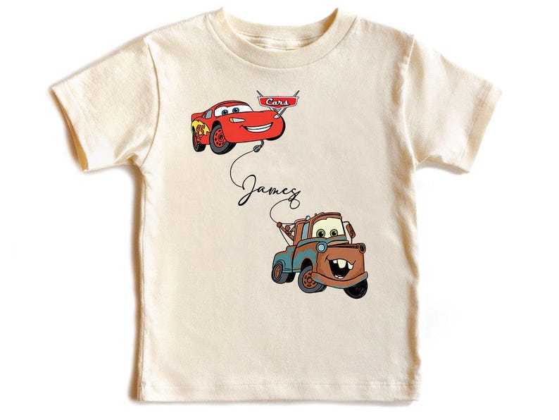 May include: Cream-colored t-shirt featuring cartoon images of Lightning McQueen and Mater from the movie Cars. The name "James" is written in cursive below the cars. The shirt has short sleeves.