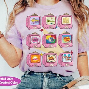 May include: A light purple t-shirt featuring a design of nine pink retro televisions. Each TV screen displays a different show title, including "Hannah Montana," "High School Musical," and "The Cheetah Girls." The text "Orchid Only With Comfort Color" is at the bottom.