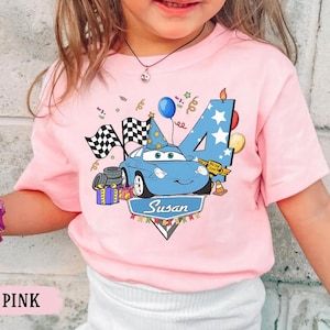 May include: Pink short-sleeved t-shirt featuring a blue cartoon car with a birthday hat, checkered flags, and the number 4. The shirt also includes a banner with the name "Susan" and birthday decorations.