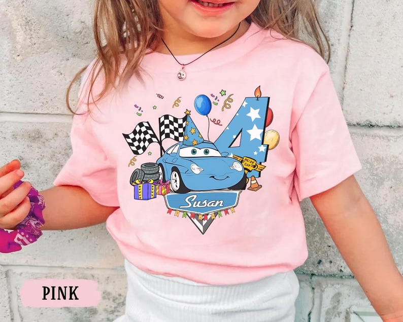 May include: Pink short-sleeved t-shirt featuring a blue cartoon car with a birthday hat, checkered flags, and the number 4. The shirt also includes a banner with the name "Susan" and birthday decorations.