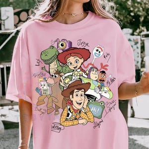 May include: A light pink t-shirt featuring a graphic print of Toy Story characters including Woody, Buzz Lightyear, Jessie, Rex, Bullseye, Forky, Mr. Potato Head, Slinky Dog, and others, with handwritten-style names and labels next to each character.