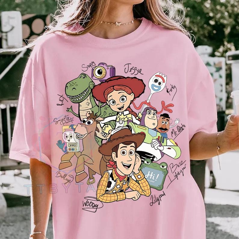 May include: A light pink t-shirt featuring a graphic print of Toy Story characters including Woody, Buzz Lightyear, Jessie, Rex, Bullseye, Forky, Mr. Potato Head, Slinky Dog, and others, with handwritten-style names and labels next to each character.