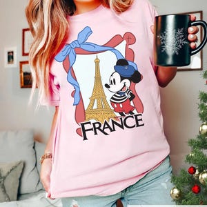 May include: A light pink t-shirt featuring a graphic of Mickey Mouse in front of the Eiffel Tower with the word "FRANCE". The design is framed with a blue ribbon. A black mug with a snowflake design is also visible.