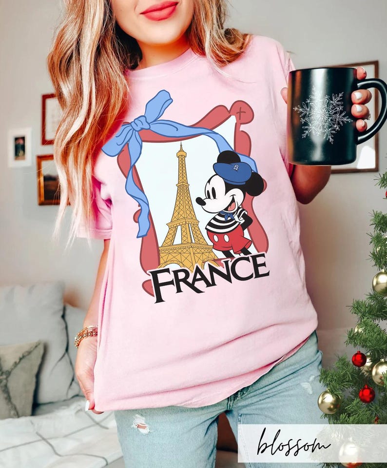 May include: A light pink t-shirt featuring a graphic of Mickey Mouse in front of the Eiffel Tower with the word "FRANCE". The design is framed with a blue ribbon. A black mug with a snowflake design is also visible.