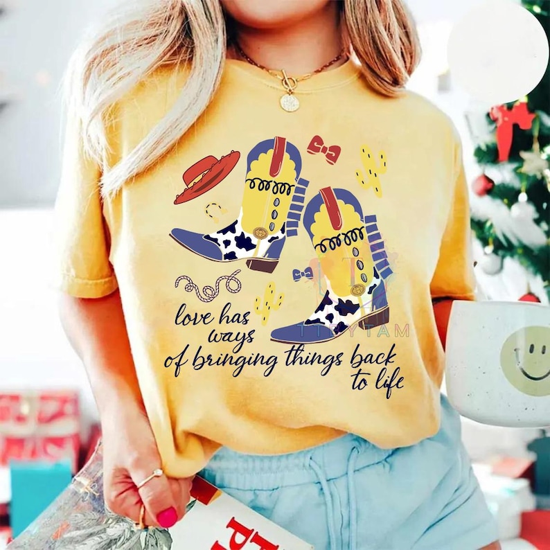 Pu&ograve; includere: Una t-shirt gialla con una stampa grafica di due stivali da cowboy con motivi a macchie di mucca gialli e neri, un cappello rosso, un piccolo fiocco e un cactus. Il testo recita "love has ways of bringing things back to life" in nero.