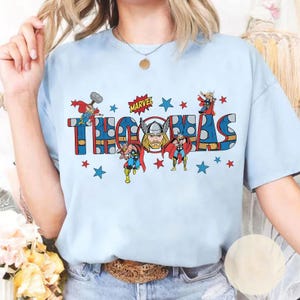 May include: Light blue t-shirt featuring the name "THOMAS" in a bold, comic book style font, with illustrations of Thor and the Marvel logo. Red and blue stars surround the text and characters.