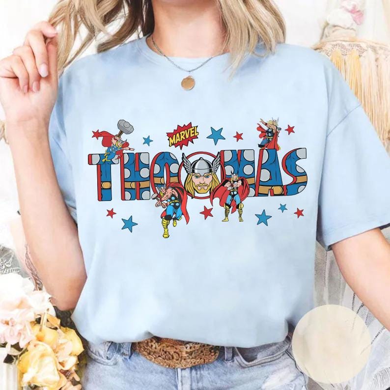 May include: Light blue t-shirt featuring the name "THOMAS" in a bold, comic book style font, with illustrations of Thor and the Marvel logo. Red and blue stars surround the text and characters.