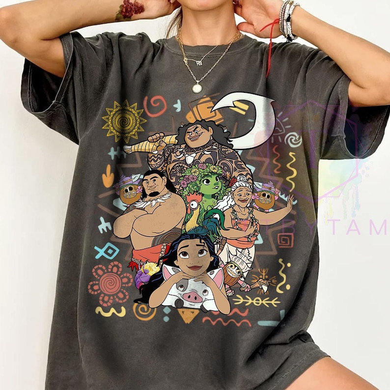 May include: Dark gray t-shirt featuring a colorful graphic of characters from the animated movie Moana. The design includes Maui, Moana, and other characters, surrounded by tribal-style patterns and symbols. The shirt has a crew neck and short sleeves.