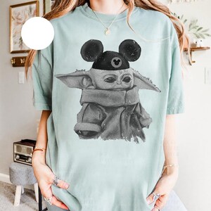 May include: A light green short-sleeve t-shirt featuring a black and white graphic print of a small creature with large ears wearing a black cap with round mouse-like ears. The graphic has a distressed, textured appearance on the fabric.