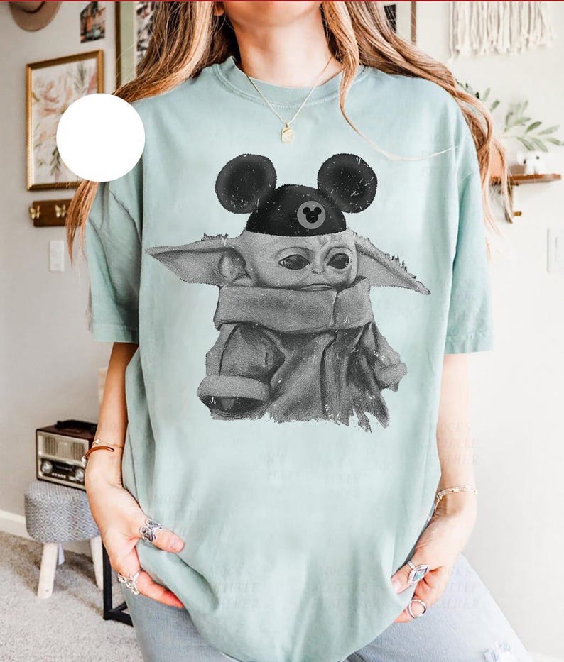 May include: A light green short-sleeve t-shirt featuring a black and white graphic print of a small creature with large ears wearing a black cap with round mouse-like ears. The graphic has a distressed, textured appearance on the fabric.