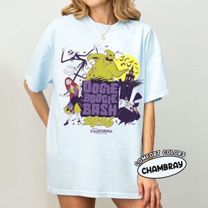 May include: Light blue t-shirt featuring a graphic design for the "Oogie Boogie Bash 2026" event. The design includes characters like Jack Skellington, Sally, and Oogie Boogie, with the text "Disney California Adventure". The shirt is labeled "Comfort Colors Chambray".