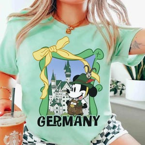 May include: A light green t-shirt featuring a cartoon Mickey Mouse in traditional German attire, framed by a green border and a yellow bow. The word "GERMANY" is printed in bold, black letters below the image. The shirt is short-sleeved.