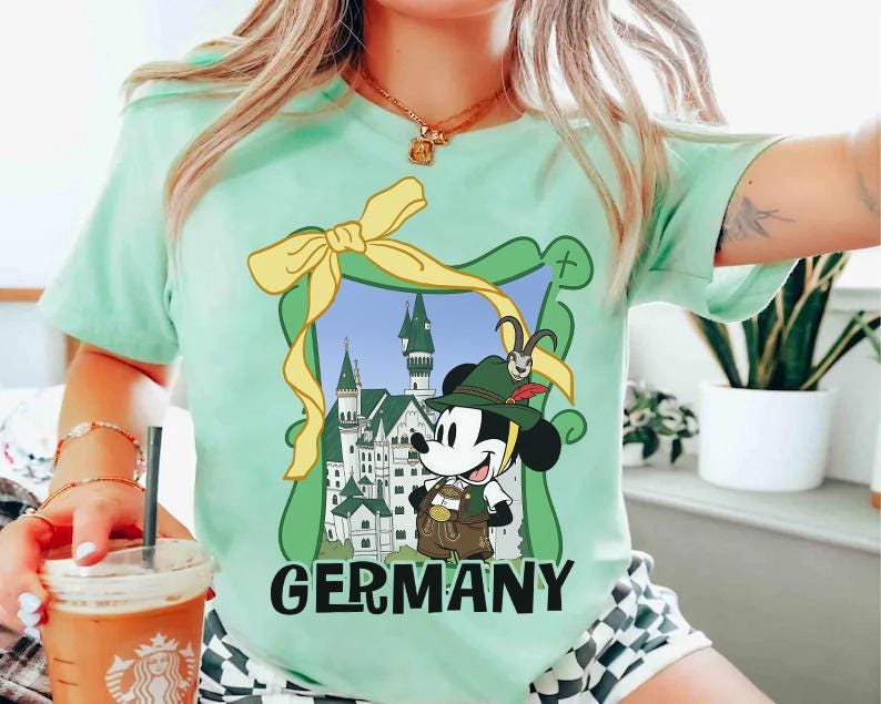 May include: A light green t-shirt featuring a cartoon Mickey Mouse in traditional German attire, framed by a green border and a yellow bow. The word "GERMANY" is printed in bold, black letters below the image. The shirt is short-sleeved.