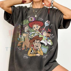 May include: A charcoal gray short-sleeve t-shirt featuring a graphic print of various animated characters including a cowboy, a cowgirl, a space ranger, a dinosaur, a horse, a spork, and a potato figure, with handwritten-style names and text.